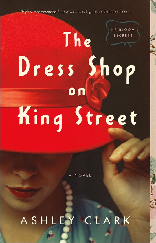 Boekcover: 'The Dress Shop on King Street' door Ashley Clark. Een vrouw met rode lippenstift verbergt een deel van haar gezicht achter een rode hoed. Parelketting zichtbaar.