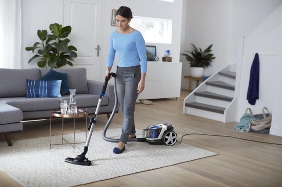 Femme en pull bleu et pantalon gris aspirateur un tapis clair dans un salon.