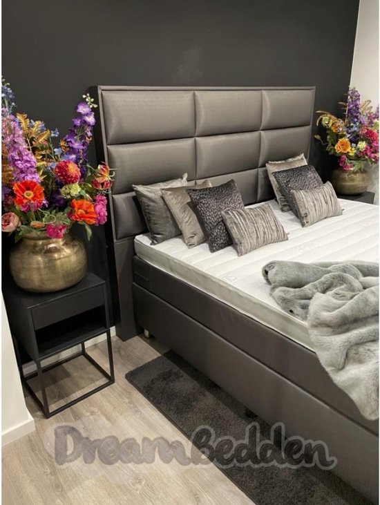 Grijze boxspring met hoog hoofdbord, witte matrassen, sierkussens, grijze deken en nachtkastje met bloemen.