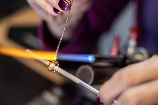 Een close-up van handen die glas bewerken met een brander. Gouden en paarse kralen worden gevormd.
