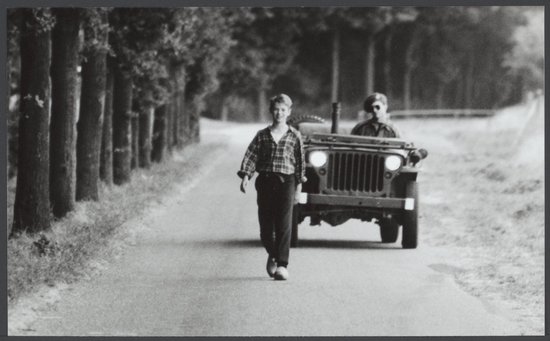 Een jongen in een geruit overhemd loopt over een asfaltweg, een jeep met een soldaat nadert van achteren.