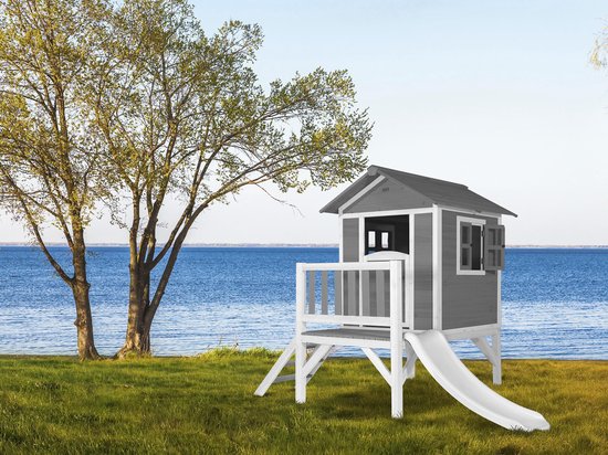 Houten speelhuisje met grijze wanden, verdieping en witte glijbaan, op gras bij een meer en bomen.