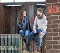 Een stel zit op een houten reling, de vrouw met een grijze sjaal, de man met een grijze muts en sjaal.