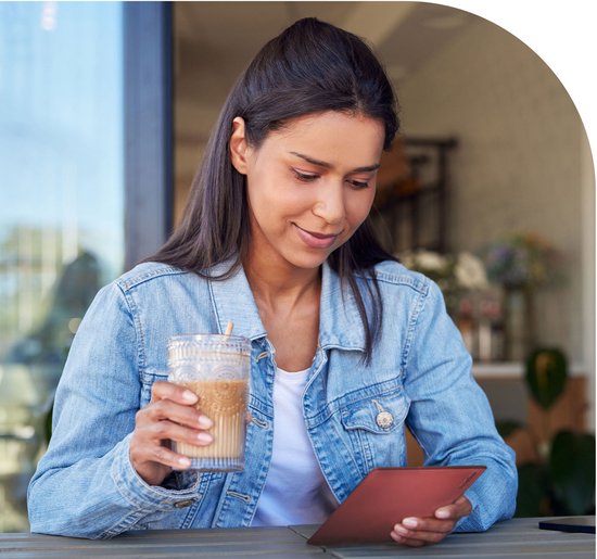 Een vrouw met een spijkerjasje zit buiten en leest op een rode PocketBook Verse Pro, met een glas koffie in haar andere hand.