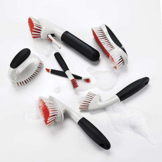Ensemble de brosses de nettoyage OXO avec poignées blanches et noires et poils rouges, dans de l'eau savonneuse.