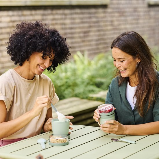 Twee vrouwen aan een tafel buiten, genietend van hun Mepal lunchpotten, gevuld met yoghurt, muesli en fruit.
