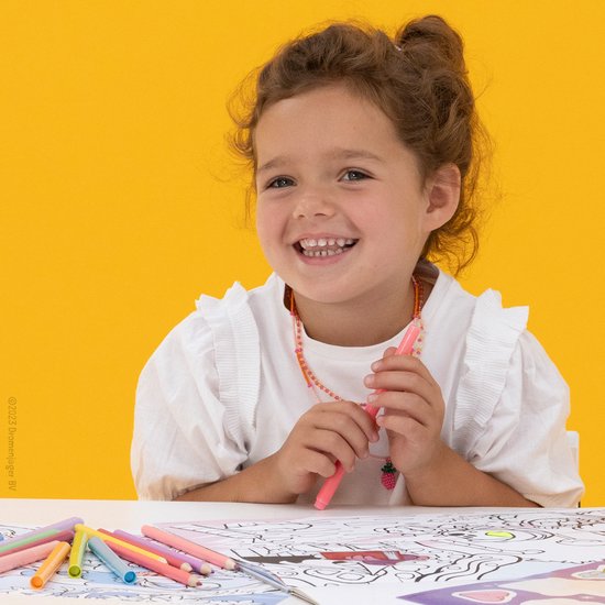 Een lachend meisje met bruin krullend haar en een roze ketting houdt een roze stift vast. Ze zit aan een tafel.
