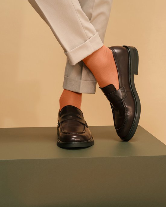 Une femme porte un mocassin marron et des collants orange, avec un pantalon gris retroussé.