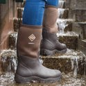 Een persoon in blauwe jeans draagt donkerbruine Muck Boot Derwent II laarzen met bruine bovenkant op natte stenen treden.