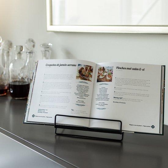 Open kookboek met recepten voor 'Croquetas de jamón serrano' en 'Pinchos met zalm & ei', op een zwarte boekenstandaard.