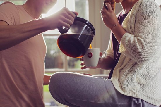 Een man schenkt koffie uit een glazen koffiekan in een mok, terwijl een vrouw erbij zit.