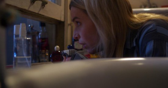Close-up van een blonde vrouw die geconcentreerd naar beneden kijkt, over een wastafel heen.