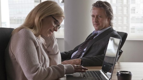 Man en vrouw zitten aan een bureau, met een laptop. De vrouw draagt een roze trui, de man een pak.