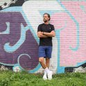 Man met gekruiste armen draagt een zwart Donnay t-shirt en blauwe short.