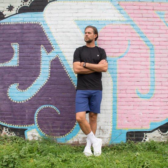 Man met gekruiste armen draagt een zwart Donnay t-shirt en blauwe short.