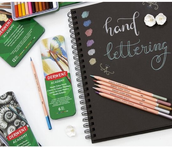 Een zwart schetsboek met 'hand lettering' in witte inkt, met 6 Derwent Academy metallic potloden. Naast het boek een blikje met 6 Derwent Academy metallic potloden.