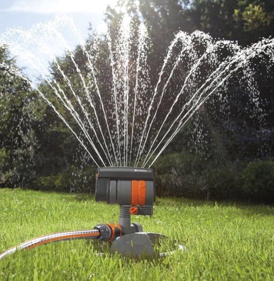 Arroseur oscillant GARDENA sur base grise, détails orange et noirs, arrose en arc sur une pelouse verte.