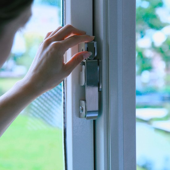 Hand sluit een zilverkleurige raamsluiting met twee grijze haken op een wit raamkozijn.