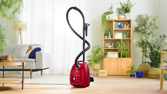 Rode Bosch stofzuiger met steel en slang, staand in een modern ingerichte woonkamer.