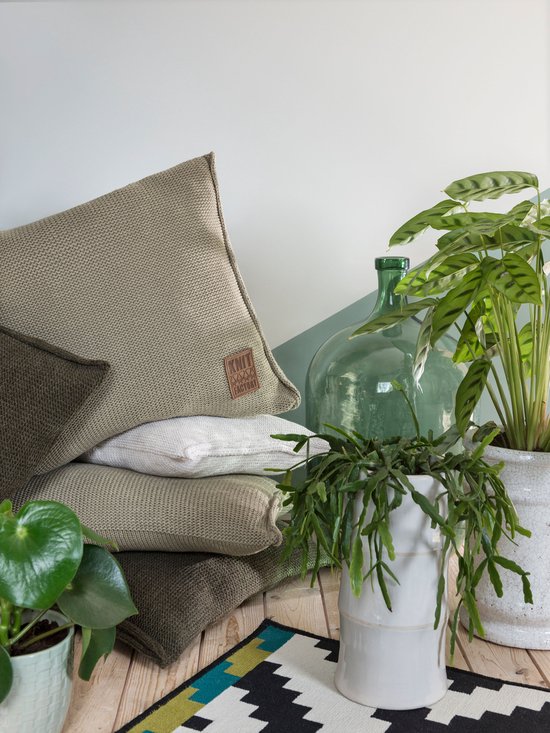 Groene kussens, een witte plaid en planten op een houten vloer met een geometrisch tapijt.