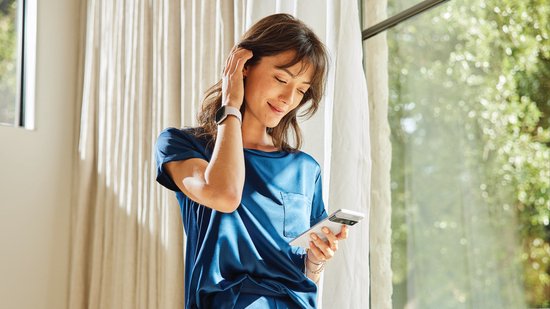 Een vrouw met een blauw shirt en een witte smartwatch staat bij een raam en kijkt op haar telefoon.