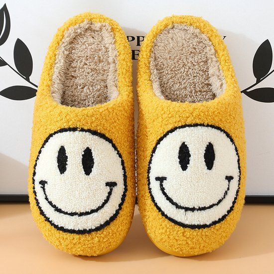 Gele fuzzy pantoffels met opdruk van een witte smiley met zwarte ogen en mond.