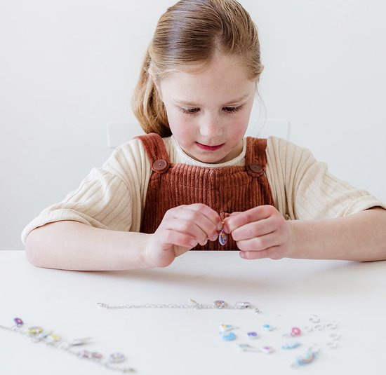 Kind in een bruine tuinbroek dat een bedel aan een ketting bevestigt, met losse bedels op de tafel.