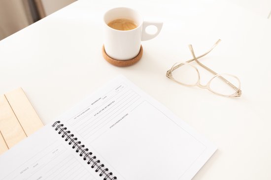 Open notitieboek, kopje koffie, bril en houten blokjes op een witte tafel.