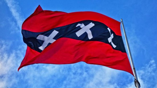 Wapperende vlag van Amsterdam met drie witte Andreaskruizen op een zwarte baan, tegen een blauwe lucht.
