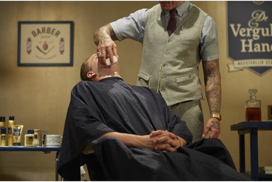 Barbier scheert een man met een scheermes, met De Vergulde Hand producten op de achtergrond.