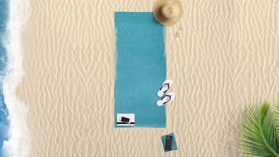 Blauw strandlaken met "BEACH" op de achtergrond, naast een strooien hoed, teenslippers en een palmblad op het zand.