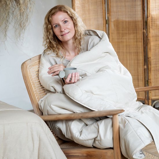 Blondine met krullen, in een beige dekbed, zittend in een houten stoel met mok koffie.