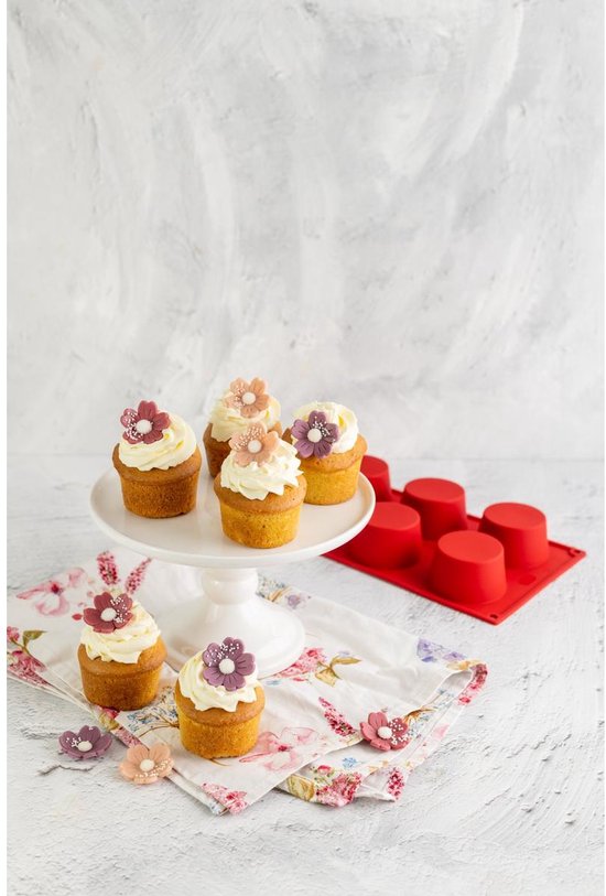 Zes cupcakes met witte crème en bloemen op een witte taartschaal en op een bloemetjesdoek, naast rode muffinvormen.