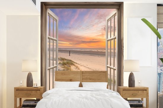 Een open raam met uitzicht op een strand bij zonsondergang, met zandduinen en een zachte, pastelkleurige hemel, boven een tweepersoonsbed.