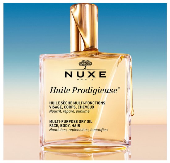 Gouden Nuxe Huile Prodigieuse multi-purpose dry oil in een glazen fles met gouden dop, op een achtergrond met blauwe lucht.
