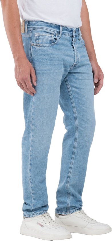 Homme en jean Replay bleu clair avec fermeture boutonnée, cinq poches et baskets blanches.