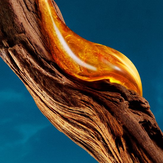 Een druppel amberkleurige olie op een stuk bruin, verweerd hout tegen een blauwe achtergrond.