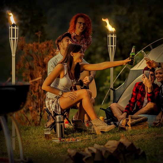 Groep vrienden kampeert 's avonds rond een tent met twee brandende bamboe tuinfakkels op de achtergrond.