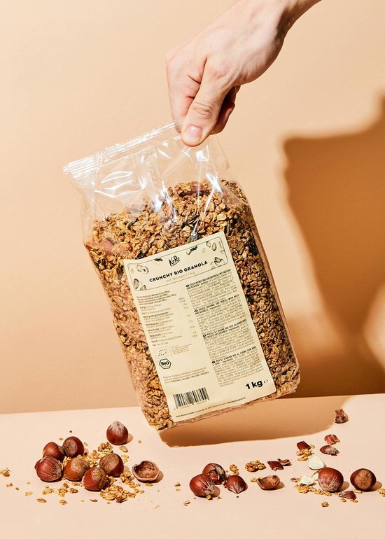 Hand die een grote zak knapperige biologische granola met noten en zaden vasthoudt, met losse noten eromheen.