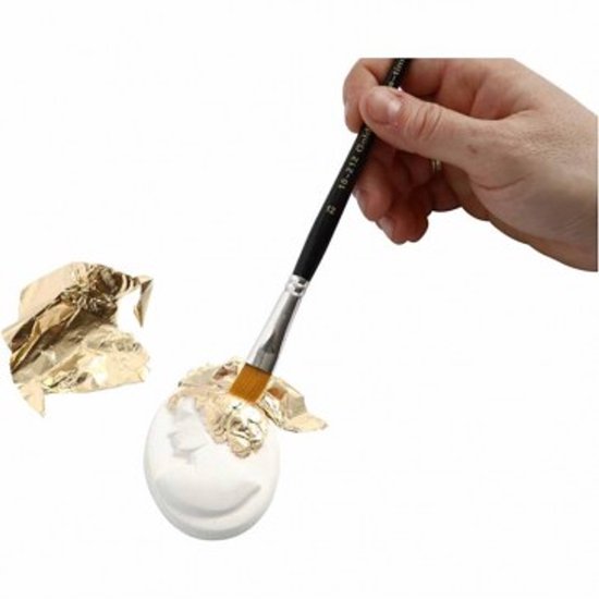 Hand met kwast brengt gouden bladmetaal aan op een wit object. Losse stukken bladmetaal liggen ernaast.