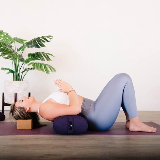 Femme sur un tapis de yoga violet, allongée avec un traversin violet sous le dos et un bloc de liège marron sous la tête.