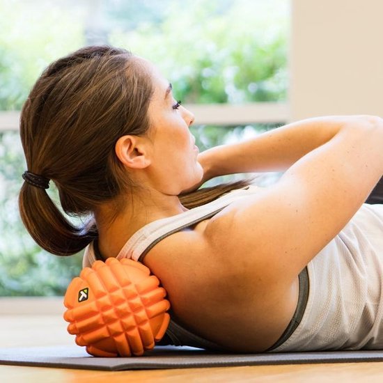 Vrouw ligt op haar rug met oranje massagebal onder haar schouder.