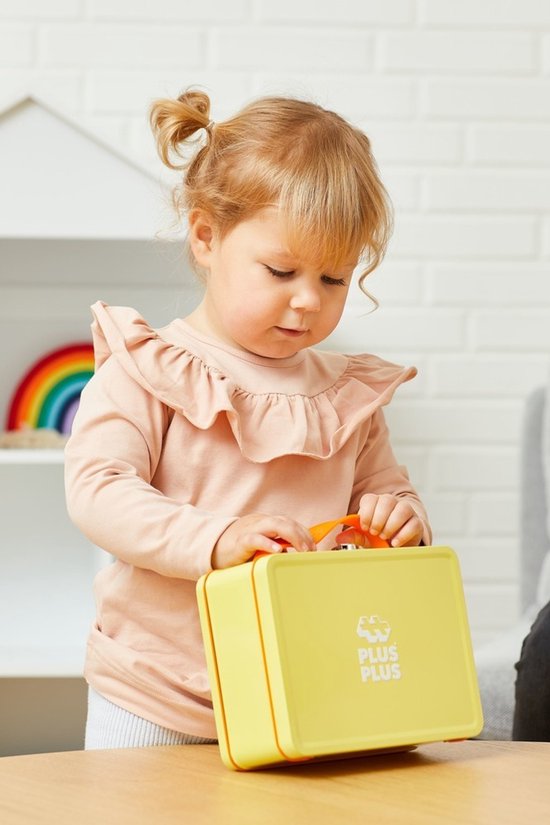 Een jong meisje met staartjes in een roze shirt sluit een gele koffer met Plus-Plus logo.