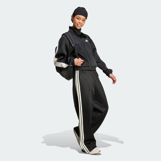 Vrouw met zwarte trainingsbroek met wijde pijpen en beige strepen, met een zwarte beanie en jas.