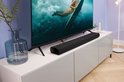 Zwarte Harman Kardon Enchant 900 soundbar op een witte tv-kast, onder een tv met een surfer op het scherm.