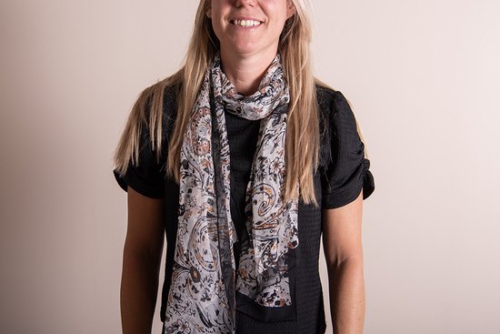 Een vrouw met blond haar draagt een zwart kledingstuk met een dunne, langere sjaal met een print van paisley en bloemen in grijs, oranje en zwart.
