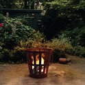 Roestbruine, metalen vuurkorf met vlammen, staand op een stenen terras in een tuin.