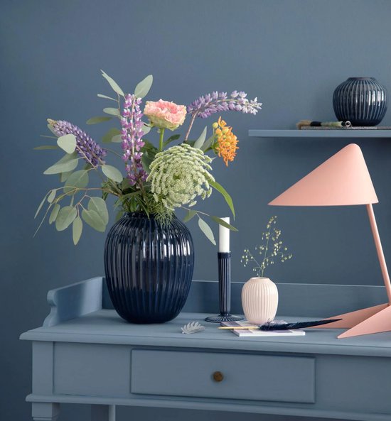 Donkerblauwe, geribbelde kandelaar naast een vaas met bloemen en een roze lamp op een blauwe bureau.
