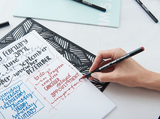 Hand schrijft met een rode kalligrafiestift op een open notitieboekje. De pagina's bevatten datums, dagen van de week en takenlijsten.