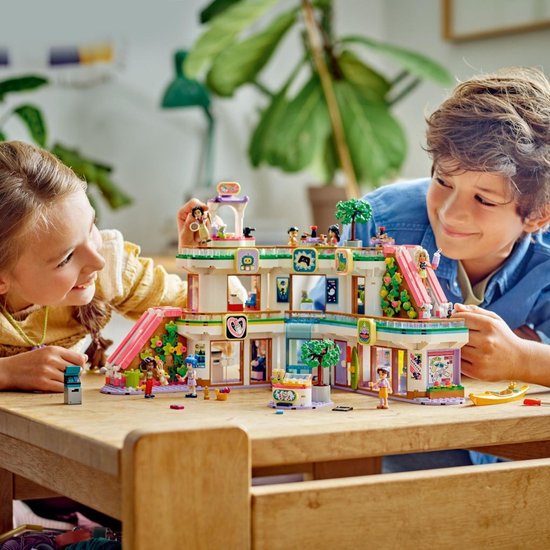 Des enfants jouent avec un set de centre commercial LEGO Friends. Construction blanche, rose et verte avec des personnages et des meubles sur une table.
